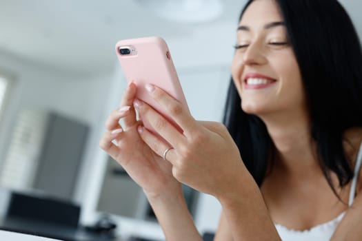 Young woman grinning happily to herself as she uses a modern pink smartphone while relaxing at home in a close up cropped low angle view with focus to the phone