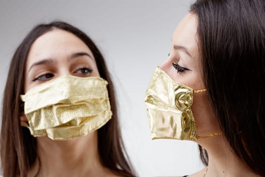 women wearing GOLDEN protective face masks staring each other, young woman good looking and lucky in golden facial covers protect from COVID-19 coronavirus on a neutral babkcround, affordable healthcare concept