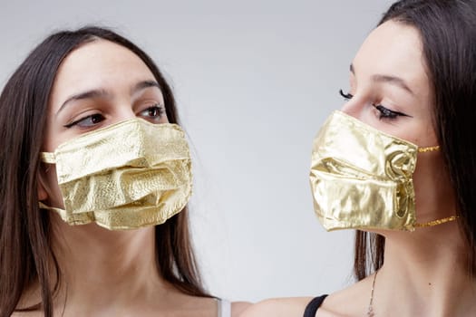 women wearing GOLDEN protective face masks staring each other, young woman good looking and lucky in golden facial covers protect from COVID-19 coronavirus on a neutral babkcround, affordable healthcare concept