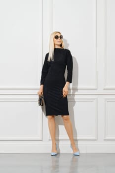 Casual office female outfit. Blonde woman in sunglasses and tight black dress standing by white wall