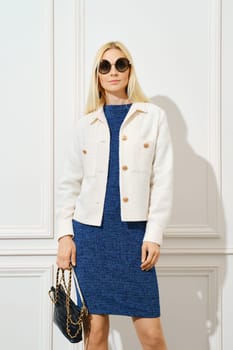 Serious woman in round sunglasses, tight blue dress and jacket holds handbag
