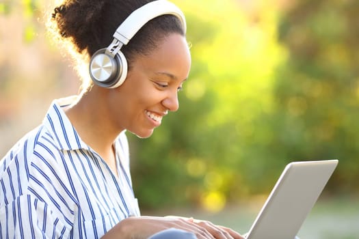 Happy black woman watching media on laptop with headphone in a green park