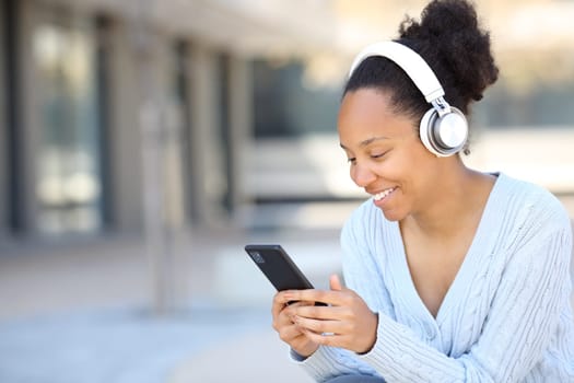 Happy black woman listening to music with headphone in the street using smart phone