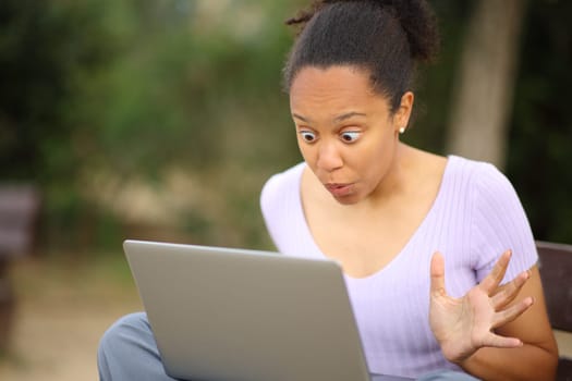 Surprised black woman checking laptop content sitting on a bench in a park
