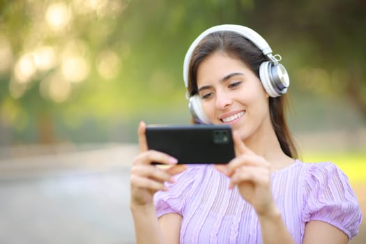 Happy woman with headphone watching videos on phone in a park
