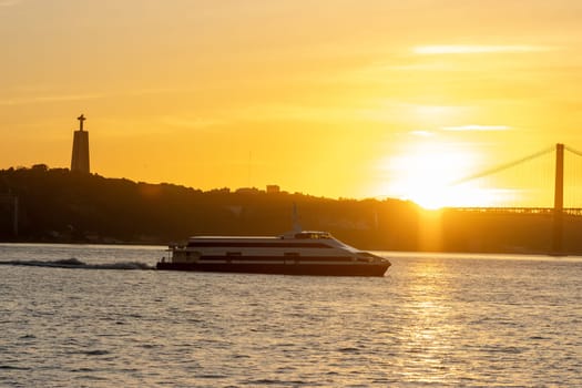 A yacht sails along the Tagus River at an orange sunset - a statue of Jesus Christ in the distance. Mid shot