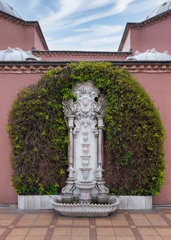 White marble fountain in Sultan Ahmed Square, Istanbul, Turkey, surrounded by lush greenery and a pink stone wall of Hagia Sophia Hurrem Sultan Bathhouse, or Ayasofya Hurrem Sultan Hamami