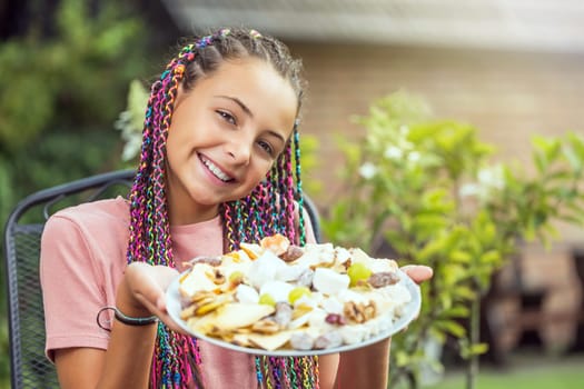 Little smiling long haired girl is holding a plate full of cheeses outdoors.  Horizontally.