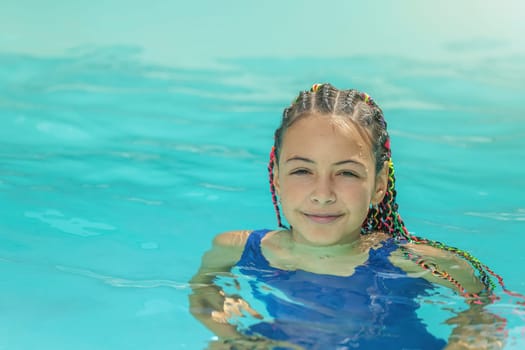 Little smiling long haired girl is in a swimsuit standing in the pool outside .Horizontally.