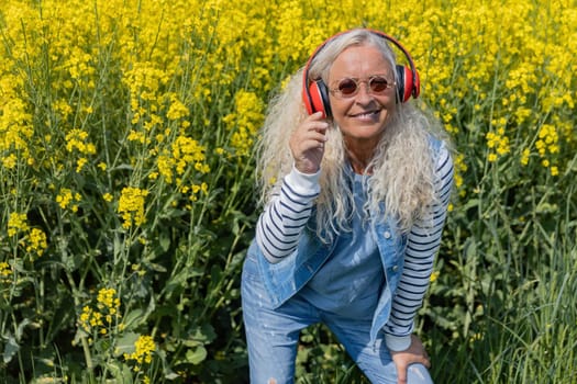 Cheerful senior woman with headphones standing beneath a rapeseed field in summertime.