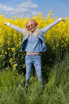 Cheerful senior woman with headphones standing beneath a rapeseed field in summertime.
