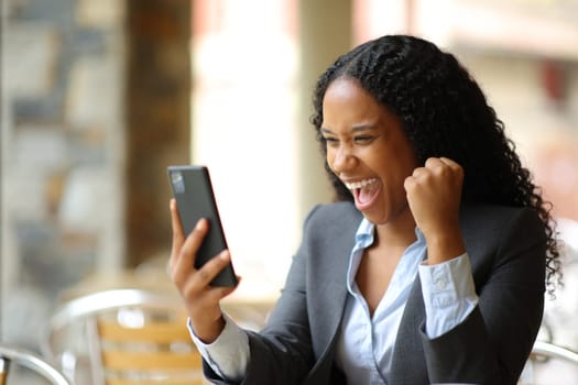 Excited black executive checking phone sitting in a bar terrace