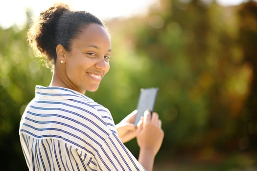 Happy black woman looks at you holding phone in a park