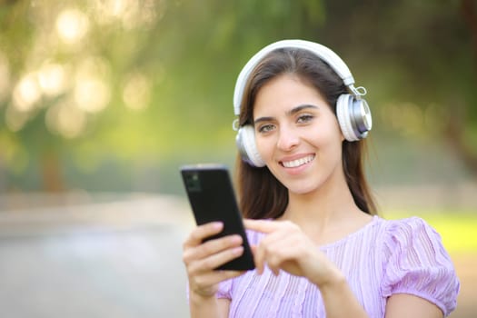 Happy female wearing headphone and holding phone listening to music looking at camera in a park