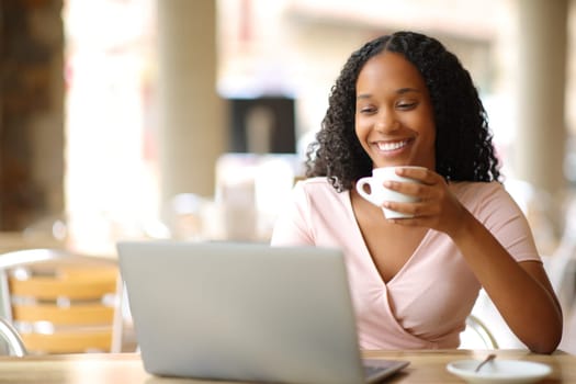 Happy black woman using laptop drinking coffee in a bar terrace