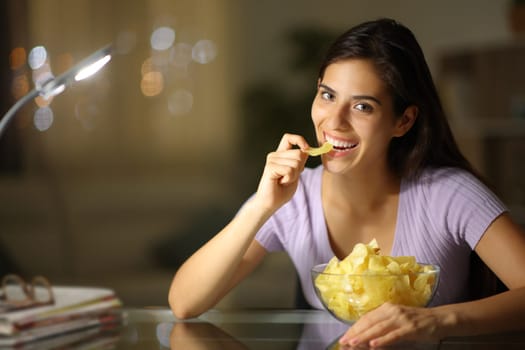 Happy woman in the night eating potato chips looking at camera at home