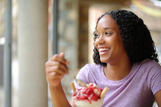 Happy black woman eating dessert looking away in a coffee shop terrace