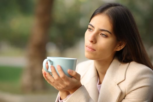 Pensive woman in winter holding coffee mug in a park