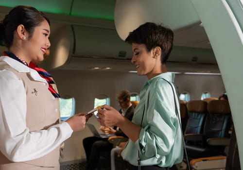 Female flight attendant in suit uniform standing at airplane entrance and checking woman passenger boarding pass for flight information