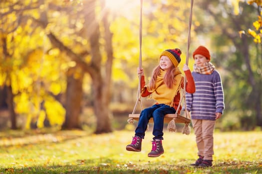 Boy swinging smiling girl on the rope swing in the autumnal park. Concept of the friendship and family holiday together.