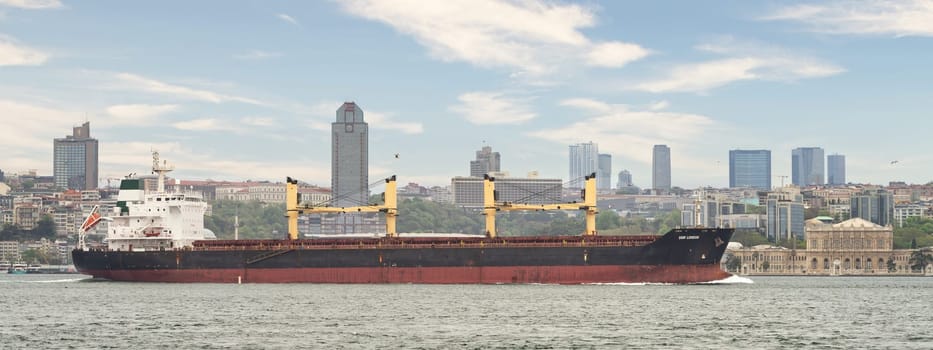 DSM London, large cargo ship sails through the Bosphorus Strait, a narrow waterway that connects the Black Sea to the Sea of Marmara. The ship is dwarfed by the towering buildings of Istanbul, Turkey