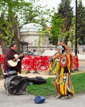 Istanbul, Turkey - May 5, 2023: Two Native Americans musicians wearing colorful traditional costume and busking playing courting flute and guitar in front of Kilic Ali Pasha Mosque in Karakoy
