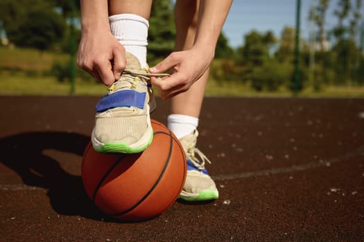 Closeup sportsman tying laces on training sneakers putting foot on ball. Basketball player preparing for training or game on street sports court