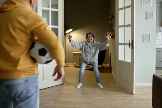 Joyful family couple of pensioner playing football at home. Selective focus on ball in hand of older man and smiling excited elderly woman ready for pass on blurred background