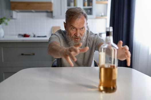 Portrait of senior man suffering from alcoholism. Elderly male sitting at kitchen table reaching to bottle of alcohol. Fight with addiction