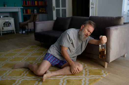 Senior man addicted to alcohol drinking behind armchair hiding from his wife. Retirement and alcoholism