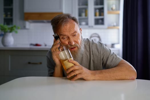 Portrait of lonely drunk man feeling depressed while sitting at table on home kitchen. Widower with alcohol addiction