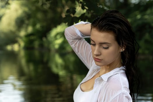 Portrait of a seductive young girl posing with wet hair in the river. The girl touches her hair with one hand. Horizontally.