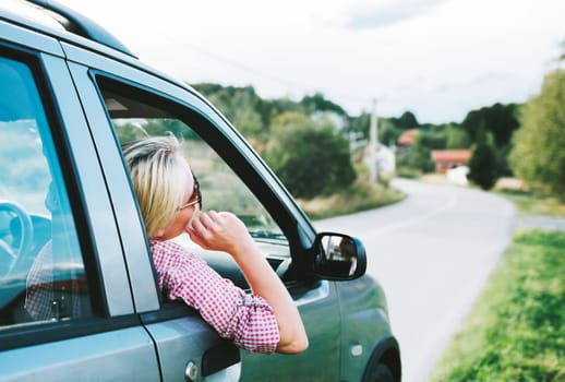 Summer holiday roadtrip travel to countryside. Young hipster blond woman driving car on rural road and having fun summer vacation. Happy girl with sunglasses arriving to mountain village.