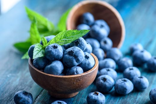 Bowl of fresh blueberries on blue rustic wooden table closeup. Colorful healthy organic seasonal fruit background. Organic food blueberries and mint leaf for healthy eating lifestyle