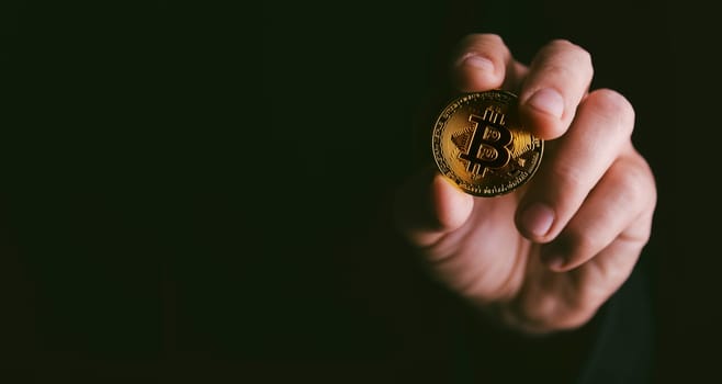 Closeup of golden bitcoin BTC cryptocurrency in hand over dark black background. Man holding golden crypto coin. Virtual money and blockchain concept.