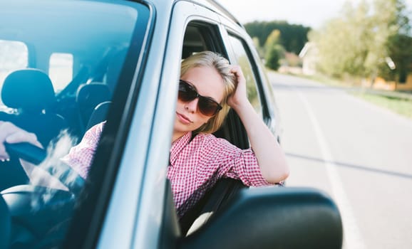 Young caucasian blond woman traveling by car and having fun on her summer holiday journey. Happy girl driving car on countryside road. Roadtrip vacation.