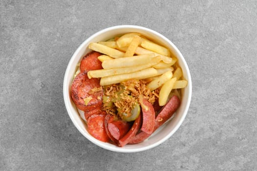 Top view of fried sausage, french fries and pickled cucumber in cardboard takeaway bowl