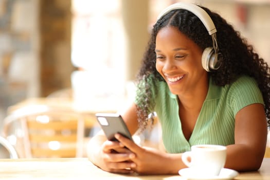 Happy black woman listening music wearing headphone using phone in a coffee shop