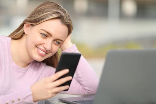 Happy teen checking smart phone content beside a laptop in the street