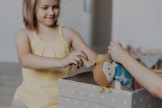 Children cleaning toys from floor after playing, sister helping to collect them doll to textile box. Kids sitting on carpet at sunny white home bedroom, kindergarten playroom or playground center