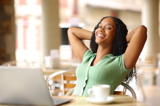 Relaxed happy black woman with laptop resting sitting in a bar terrace looking at you