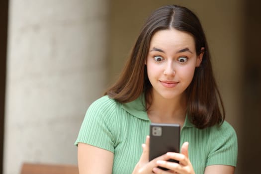 Front view portrait of a shocked woman checking cell phone in the street