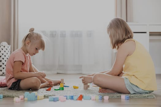 Children play with colorful plastic building blocks. Kids sitting on floor indoors at home bedroom, kindergarten, nursery. Educational eco-friendly child game. Cute little sisters having fun together