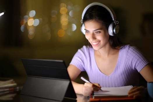 Happy student using tablet and headphone and taking notes in the night at home