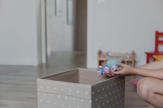 Child cleaning toys from floor after playing, collect them to textile box. Kid sitting on carpet at white home bedroom, kindergarten playroom or playground center. Educational eco-friendly games