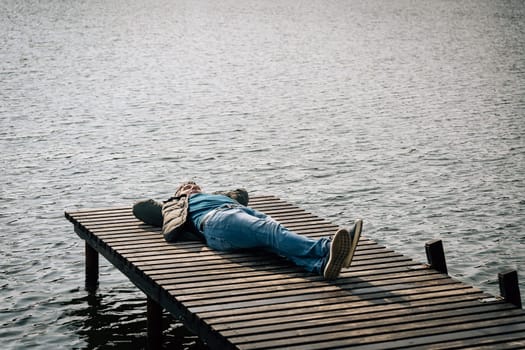 A man in jeans, a jumper and a jacket lies relaxing on a wooden boat dock on a lake and enjoying a sunny autumn day.