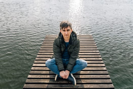A man sits relaxed on a wooden pier on the lake, cross-legged in jeans and a jacket in autumn.