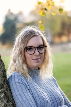 Portrait of blonde young woman in glasses posing leaning against a autumn tree.