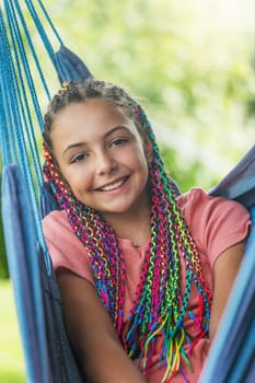Laughing young girl with colorful braids in her hair is posing on hammock. Vertically.
