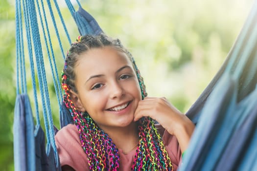 Closeup portrait of laughing young girl with colorful braids in her hair posing on blue hammock.  Horizontally.